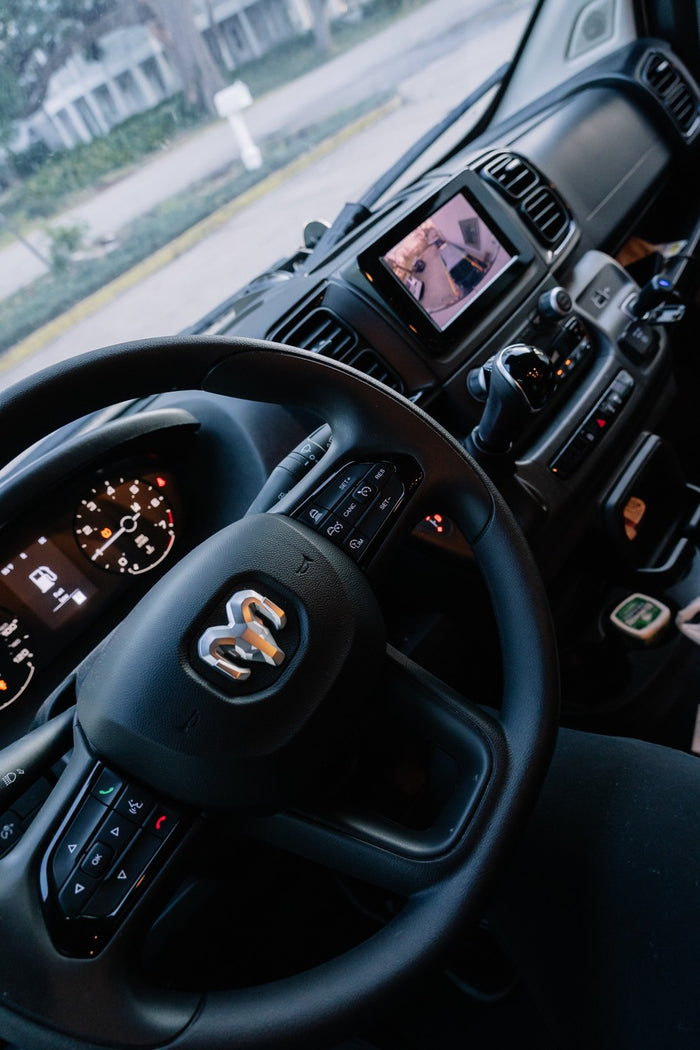 Inside a modern RV rental van: steering wheel with Ram logo, dashboard, and infotainment screen displaying a camera view.