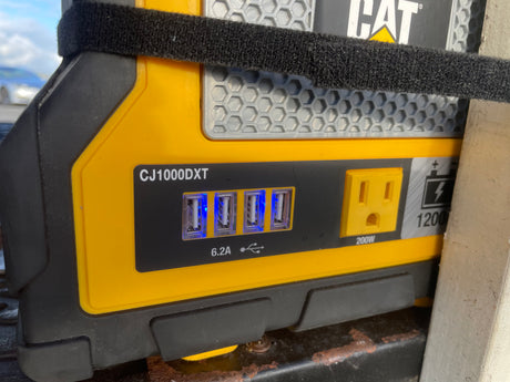 A yellow CAT CJ1000DXT portable power station with 4 glowing USB ports and AC outlet, useful for RV rental power & jump-starting.