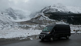 Scenic winter landscape with a black RV rental van parked on a wet road amidst majestic, snow-covered mountains.