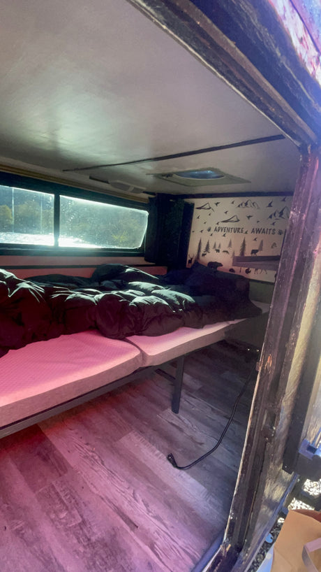 Cozy rv rental interior with an "Adventure Awaits" decal, comfortable bed, and a bright window view, perfect for rest.