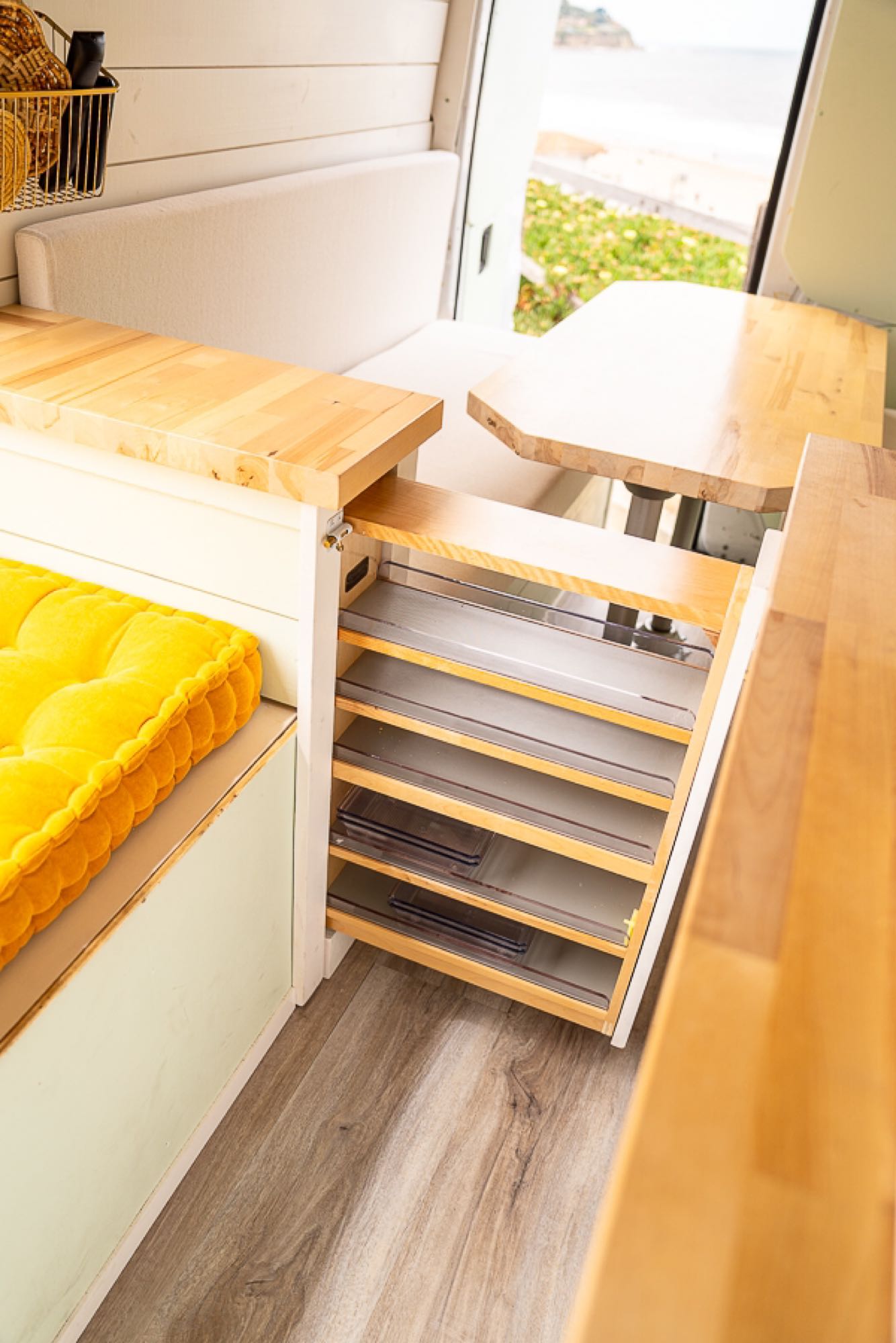 RV rental: Inside a modern camper van with pull-out storage, wooden counters, a comfy bench, and an ocean view.