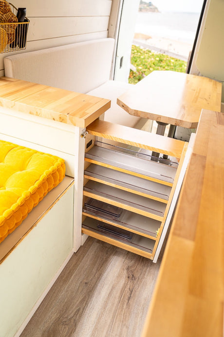 RV rental: Inside a modern camper van with pull-out storage, wooden counters, a comfy bench, and an ocean view.