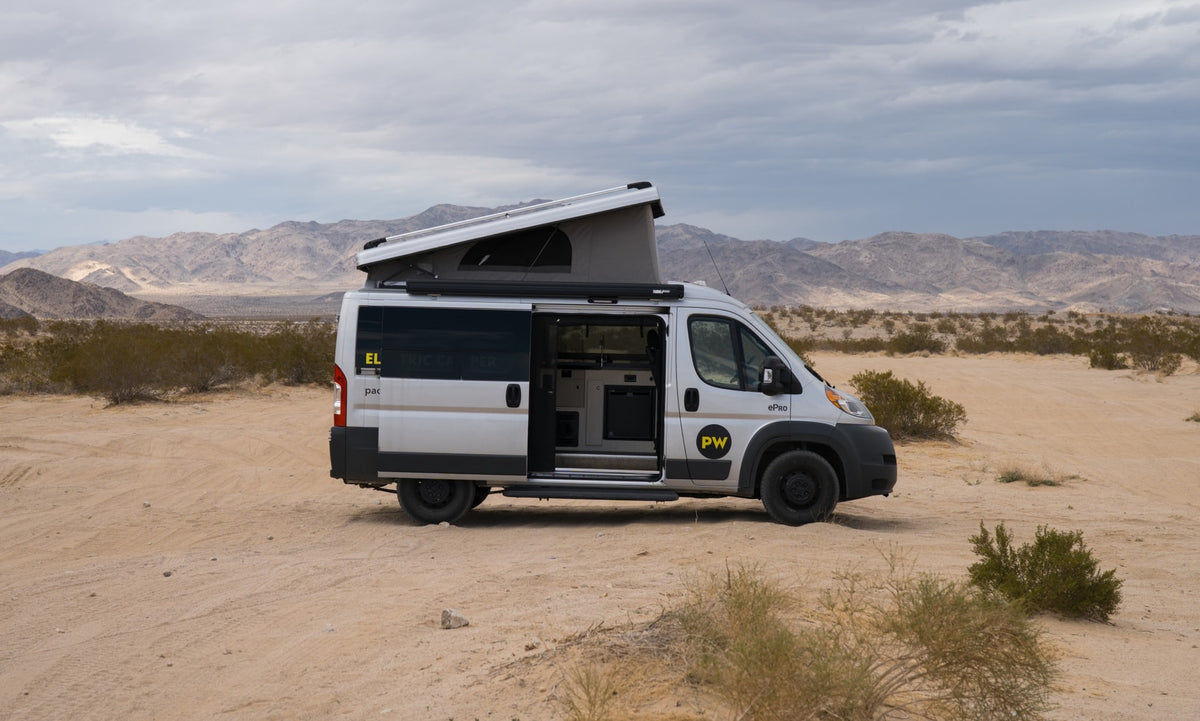 This electric RV rental camper van, pop-top open, is parked in a sandy desert with distant mountains under a cloudy sky.
