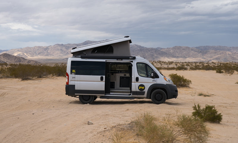 This electric RV rental camper van, pop-top open, is parked in a sandy desert with distant mountains under a cloudy sky. — photo 1