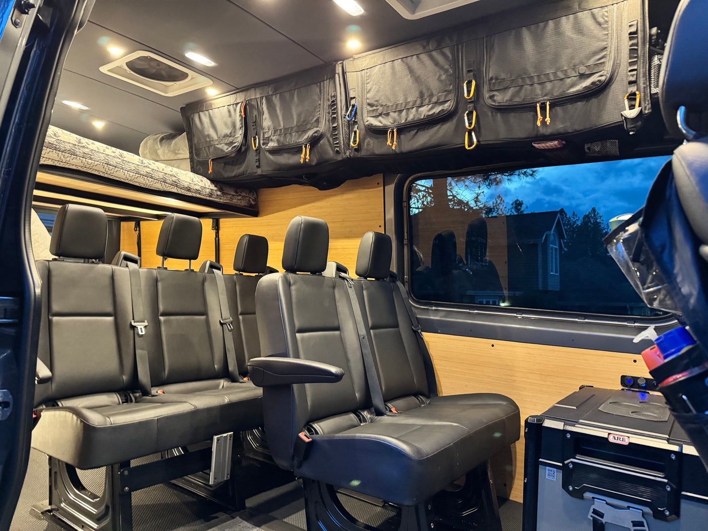 Inside a custom RV rental camper van, showing comfortable passenger seating, overhead storage, and a raised bed.