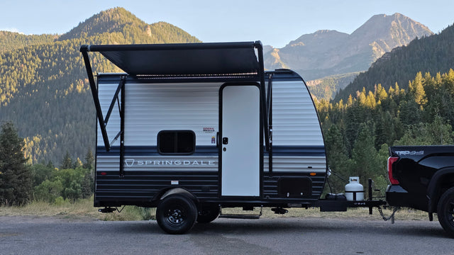 This Springdale travel trailer rv rental, hitched to a pickup truck, is parked amidst majestic mountains, ready for outdoor adventures.