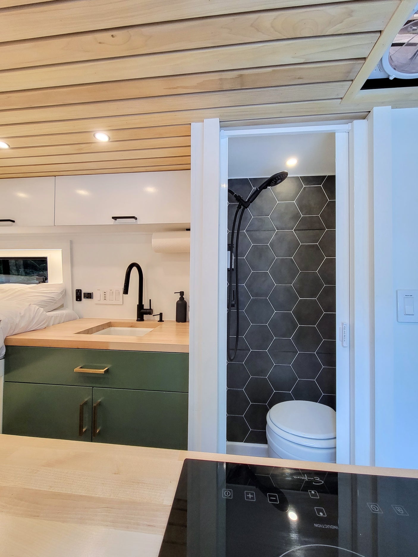 Modern RV rental interior with a compact kitchen featuring green cabinets, wood counter & sink, alongside a black tiled shower & toilet.