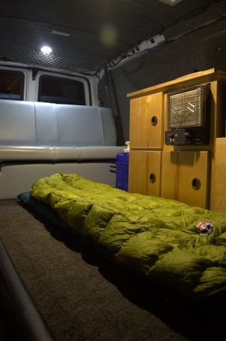 Cozy RV rental interior shows a green sleeping bag on a carpeted platform, heater, and bench seat. Ready for camping. — photo 12
