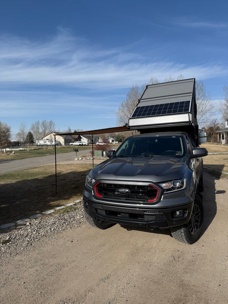 This grey Ford truck features an RV rental-style pop-up camper with solar panels and an awning, ready for overlanding.