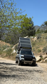 An overland-equipped Land Rover Defender, fitted with solar panels & rooftop tent, on a dirt road. Perfect for adventure, like an RV rental for the wild.