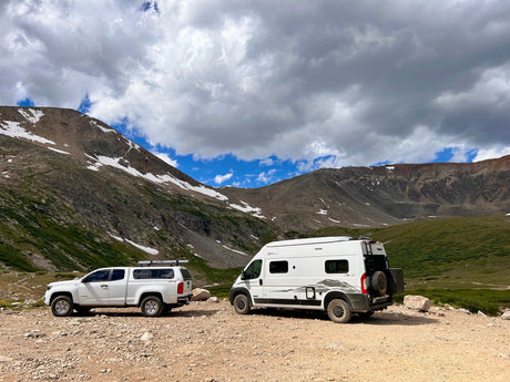 An RV rental camper van & pickup truck explore a rugged mountain road with snow. Ideal for outdoor adventures!