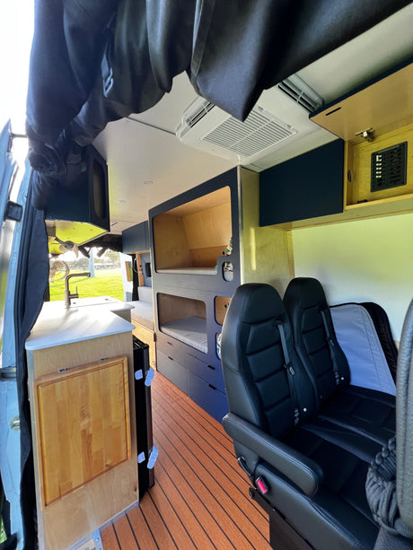Interior of a custom rv rental van with a kitchen, bunk beds, seating, wood-slat flooring, and an overhead AC unit.