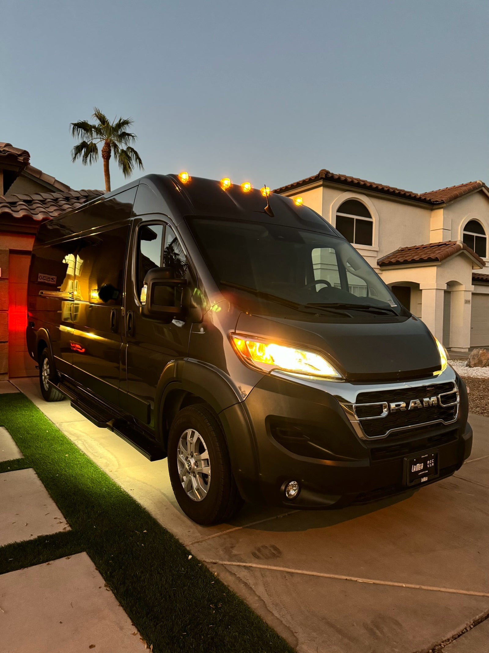 A dark Ram Promaster camper van, ready for RV rental, parked with lights on in a home driveway at dusk.