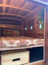 Rustic RV rental interior: wood-paneled walls & ceiling, cozy bed with patterned linens, porthole windows, and a plant.