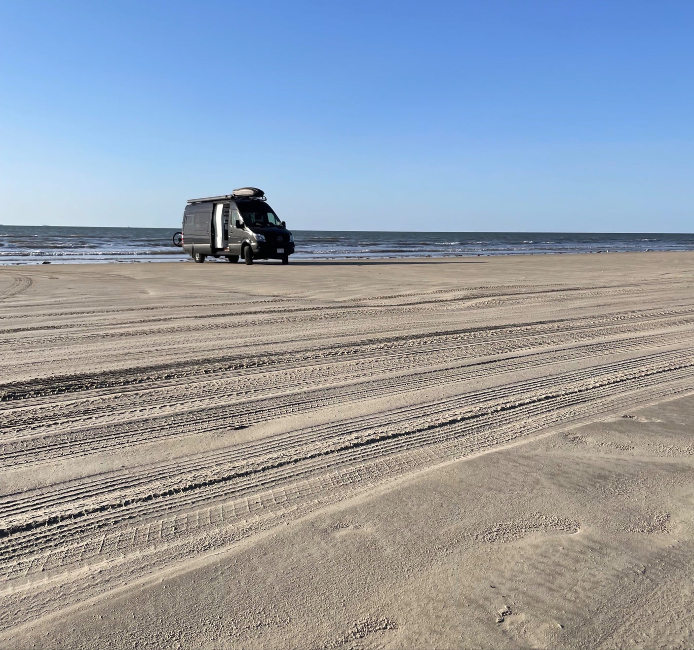 Experience beach life: a grey camper van, ideal for rv rental, on a sandy beach with ocean waves under a clear sky.