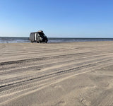 Experience beach life: a grey camper van, ideal for rv rental, on a sandy beach with ocean waves under a clear sky.