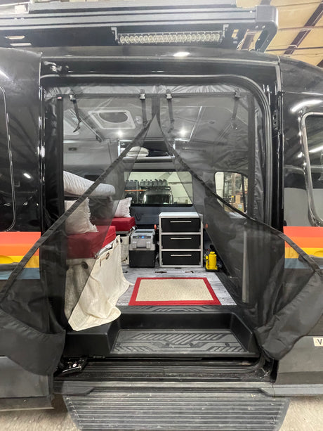 Interior view of a customized RV rental, showing a bed, storage drawers, fridge, and protective bug screen.
