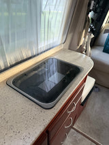 RV rental kitchen interior: white curtains, speckled countertop, Dometic cooktop, and wood cabinets.