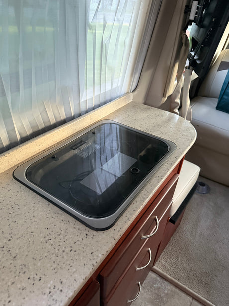 RV rental kitchen interior: white curtains, speckled countertop, Dometic cooktop, and wood cabinets.