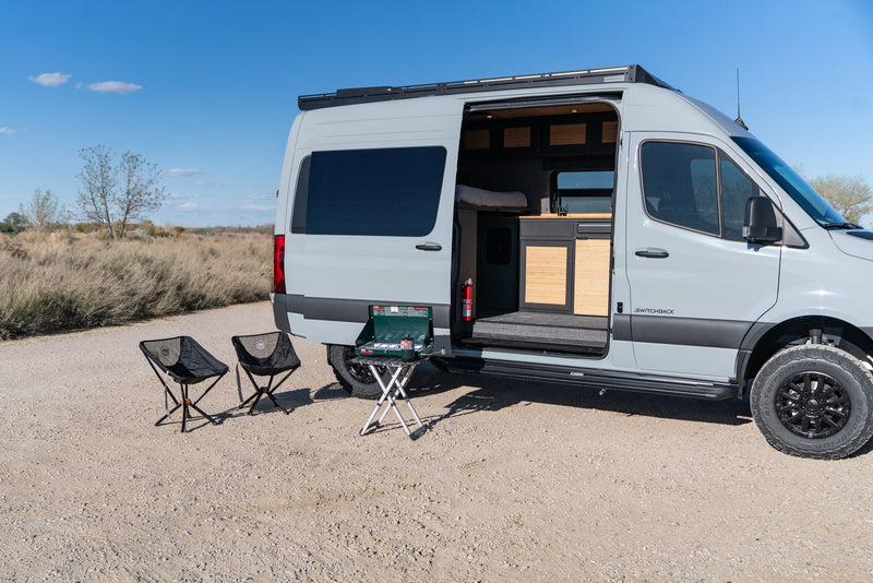 This rv rental shows an open camper van with kitchen, bed, and outdoor cooking setup. Two chairs sit on a dirt road in nature. — photo 10