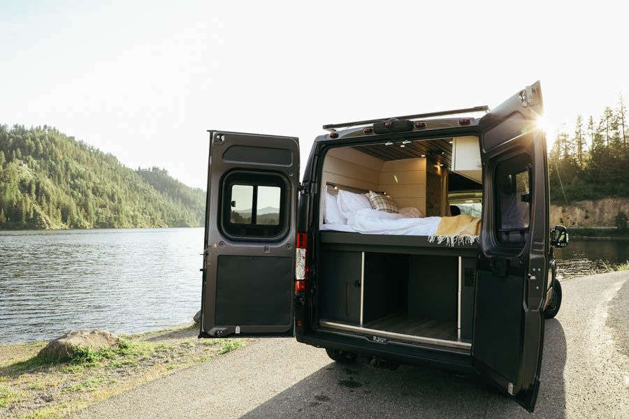 RV rental parked lakeside. Open doors reveal a cozy bed & storage inside, offering scenic views for travel.