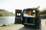 RV rental parked lakeside. Open doors reveal a cozy bed & storage inside, offering scenic views for travel.
