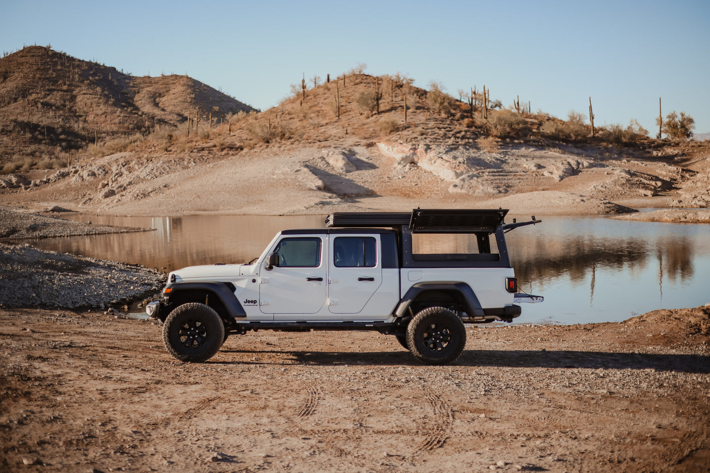 White Jeep Gladiator overlanding RV rental with open rear canopy by a desert lake with saguaro cacti.