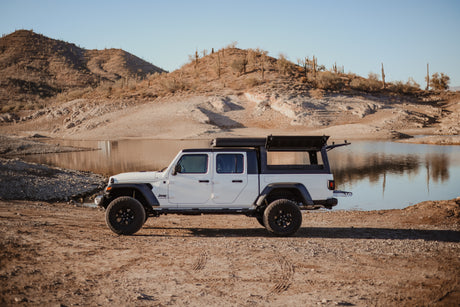 White Jeep Gladiator overlanding RV rental with open rear canopy by a desert lake with saguaro cacti.