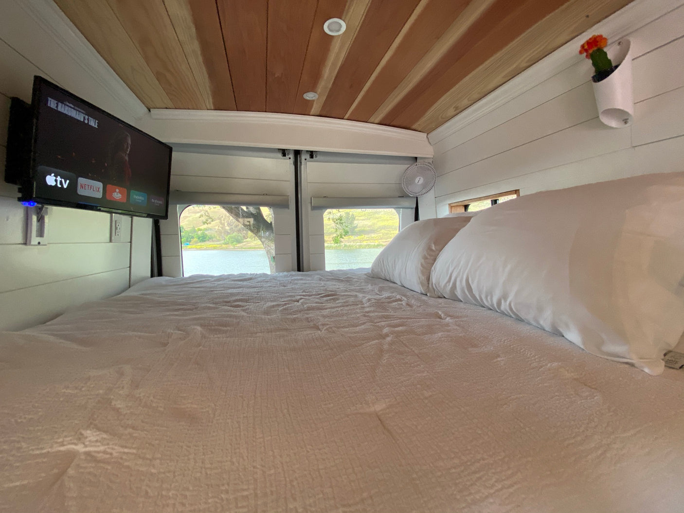 An RV rental's inviting interior: bed, TV showing Netflix, wood ceiling, and scenic lake views outside.