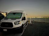 White Ford Transit RV rental at a waterfront campsite (site 55) with city lights across the bay. Windshield frosted.
