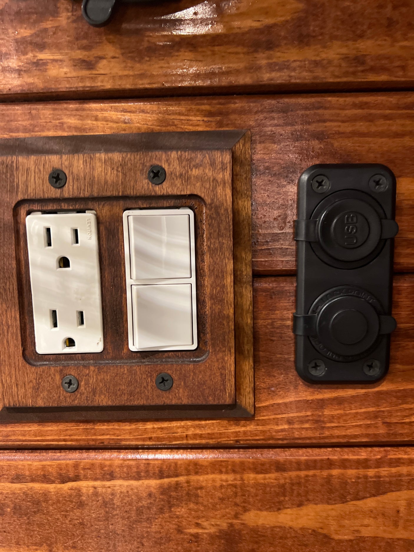 RV rental utility panel: white power outlet, two light switches, and black USB/12V charging ports on stained wood.