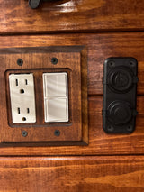 RV rental utility panel: white power outlet, two light switches, and black USB/12V charging ports on stained wood.