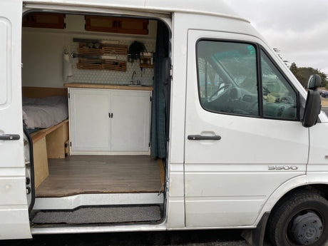 White Sprinter camper van for RV rental, showing interior kitchenette, bed, and driver's area.