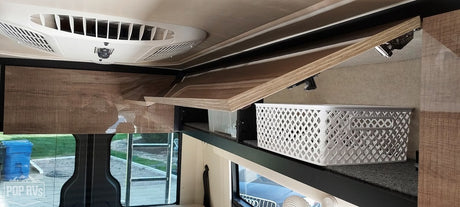 Inside an RV rental: overhead AC unit, open wood-grain cabinet with white storage basket, and outdoor view.