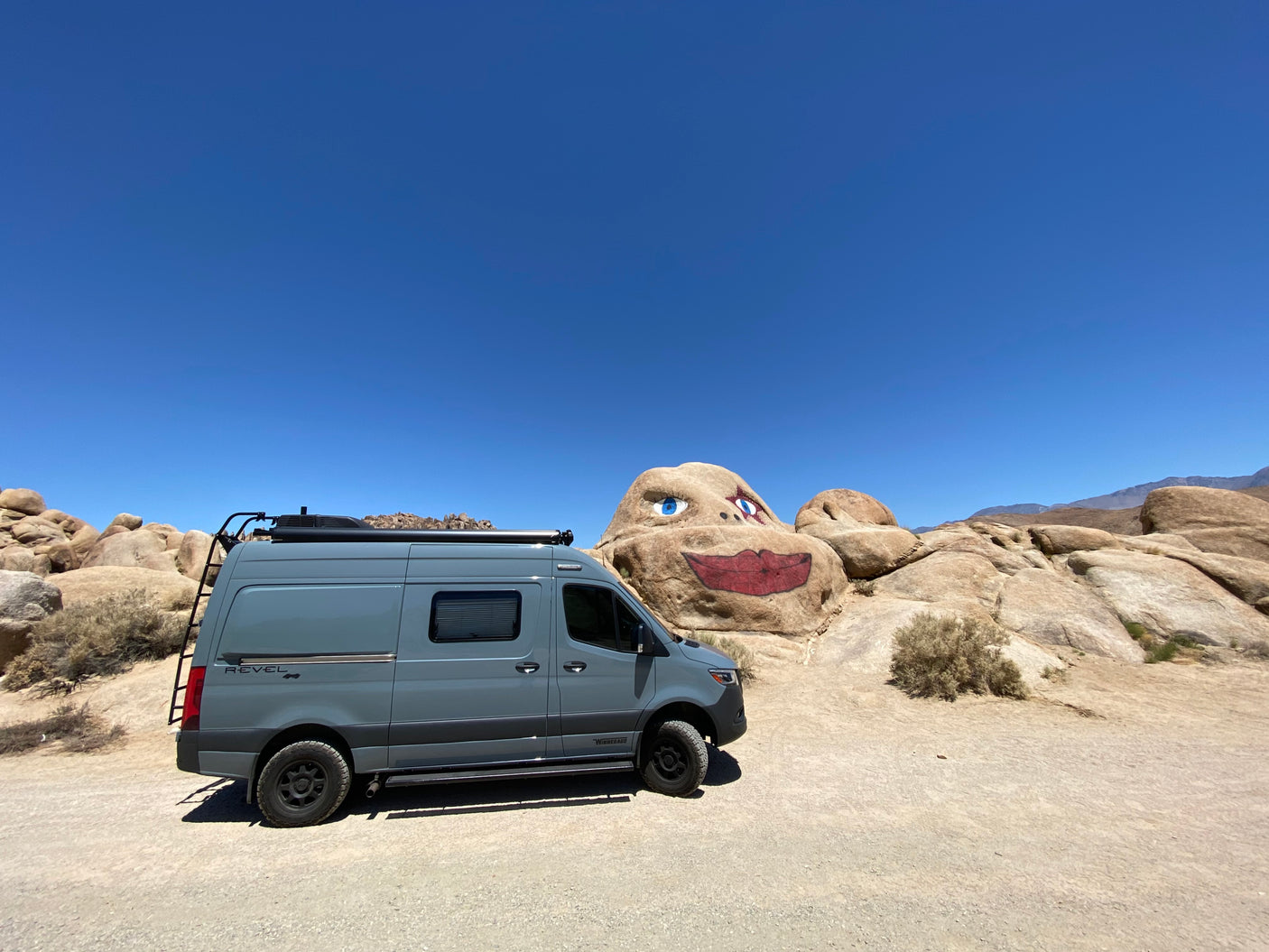 This grey Winnebago Revel 4x4 rv rental is parked on dirt, showcasing a unique painted rock face in the desert.