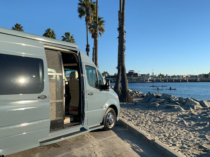  with a camera on the roof.|RV rental's advanced backup camera system shows parking sensors and a clear rear view of the harbor.|Light gray RV rental van parked on a sunny sandy bay with palm trees & kayakers. Ideal for beach travel adventures.|RV rental digital control panel & battery monitor display tank levels — photo 13
