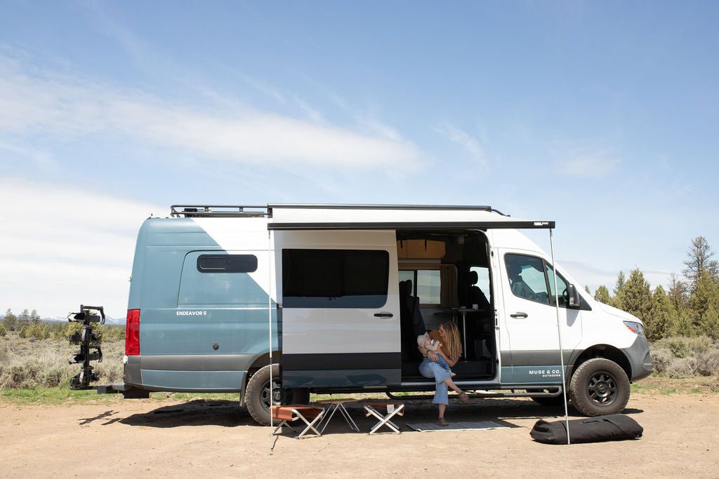 Enjoying the outdoors in an rv rental camper van. A woman holds a baby by the open door of the two-tone blue/white van with an awning.