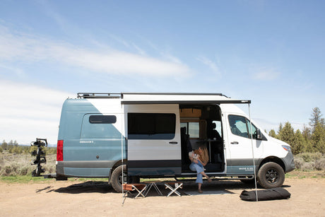 Enjoying the outdoors in an rv rental camper van. A woman holds a baby by the open door of the two-tone blue/white van with an awning.