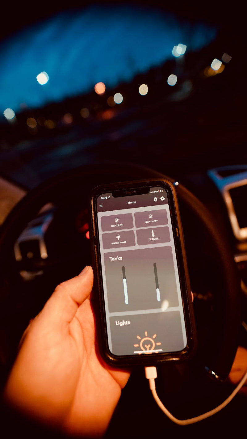 A hand holds a smartphone in an RV at night. The screen shows an RV rental control app for lights, water, climate, and tank levels. — photo 9