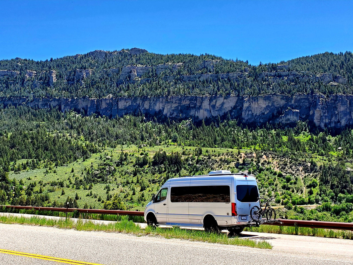 Silver RV rental van with bikes parked on a mountain road, against dramatic forested cliffs.