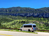 Silver RV rental van with bikes parked on a mountain road, against dramatic forested cliffs.