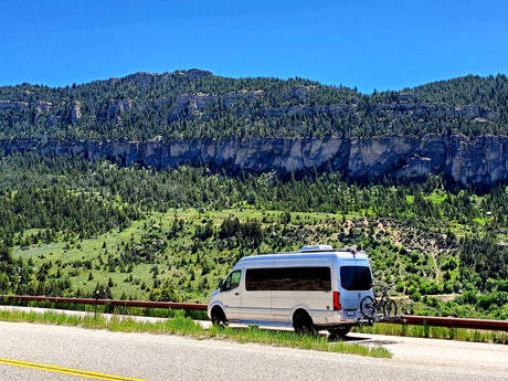 Silver RV rental van with bikes parked on a mountain road, against dramatic forested cliffs.