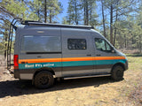 Gray rv rental camper van with orange & teal stripes parked in a sunny pine forest, ready for adventure.
