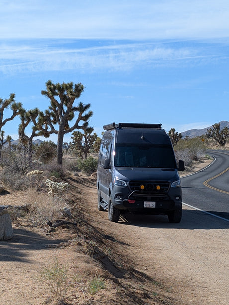 An RV rental Sprinter camper van on a dirt shoulder by a winding desert road, with Joshua trees and mountains under a blue sky.