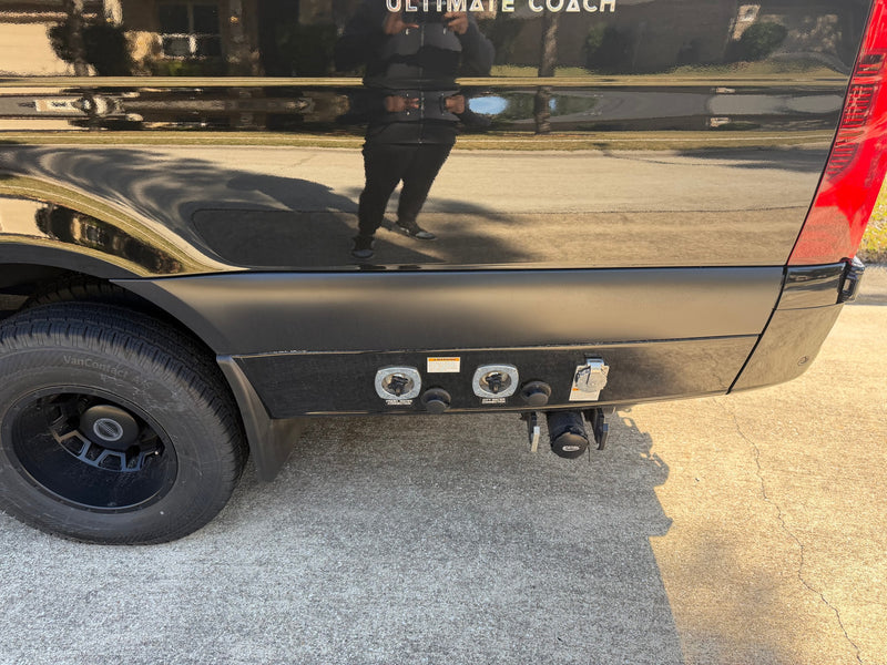 Rear of a black RV rental van, showing water, power, and sewer utility connections and a tire. A person is reflected on the glossy surface. — photo 7