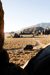 Experience the wild with an RV rental! A black camper van sits on a dirt road amidst rocky desert terrain & mountains.