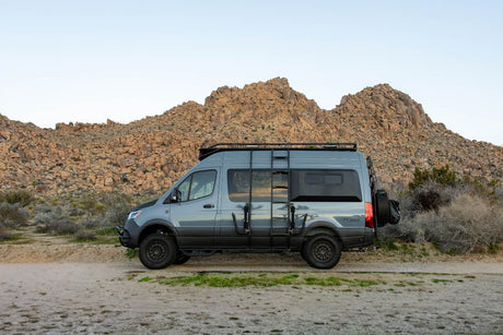 Off-road RV rental: a grey Sprinter adventure van with custom gear, parked on a dirt path in a rocky desert landscape.
