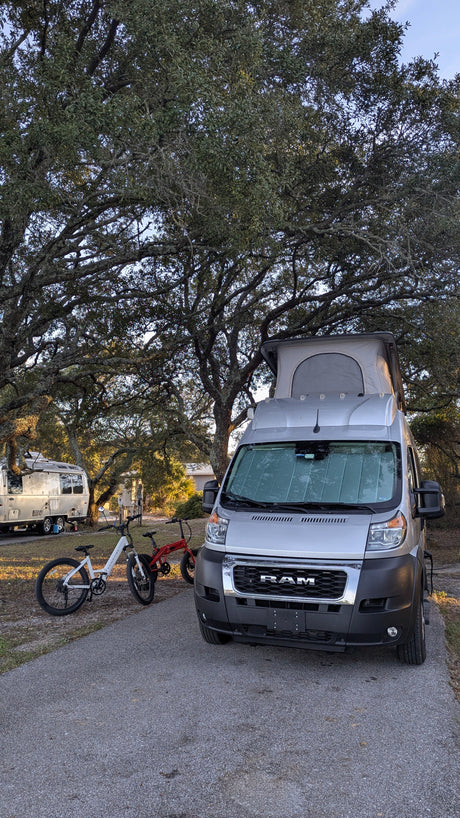 This RV rental camper van with a pop-top roof and two electric bikes is ready for adventure at a shady campsite.