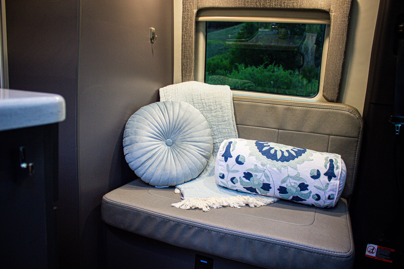 RV rental interior: A plush gray bench with light blue and patterned pillows, a soft throw, and a window showcasing lush greenery.