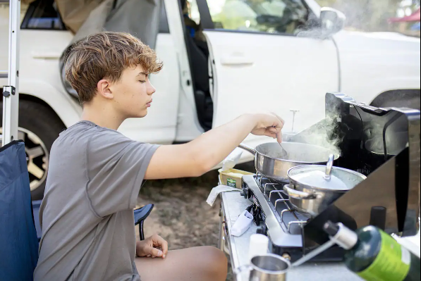 A young person cooks on an outdoor stove, steam rising from pots, with an RV rental vehicle visible behind. — photo 10